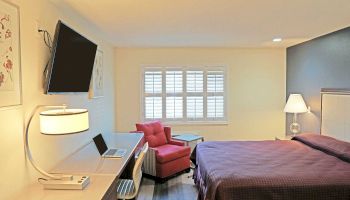 This image shows a modern hotel room with a wall-mounted TV, a work desk with a laptop, a cozy chair by the window, and a bed with purple bedding.