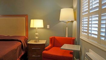 A cozy bedroom corner with a bed, two lamps, an orange chair, and a small table by shuttered windows is shown.