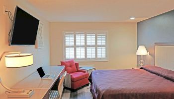 A well-lit bedroom features a mounted TV, a comfortable bed, a desk with a laptop and lamp, a red armchair, and a window with shutters.