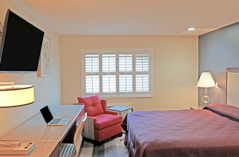 A well-lit bedroom features a mounted TV, a comfortable bed, a desk with a laptop and lamp, a red armchair, and a window with shutters.