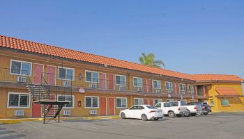 A two-story motel with exterior entrances, parking lot, several parked cars, and palm trees in the background under a clear sky.