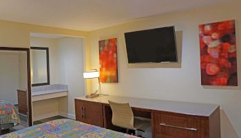 This image shows a hotel room featuring a bed, a desk with a chair, a wall-mounted TV, abstract artwork, a mirror, and a bathroom vanity.