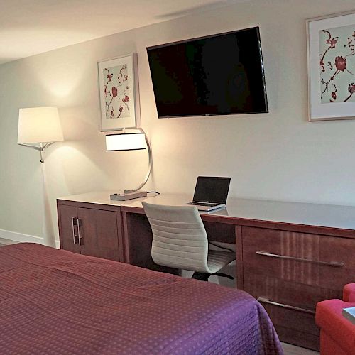 A hotel room featuring a bed, a desk with a laptop, a wall-mounted TV, a red armchair, and two framed pictures.