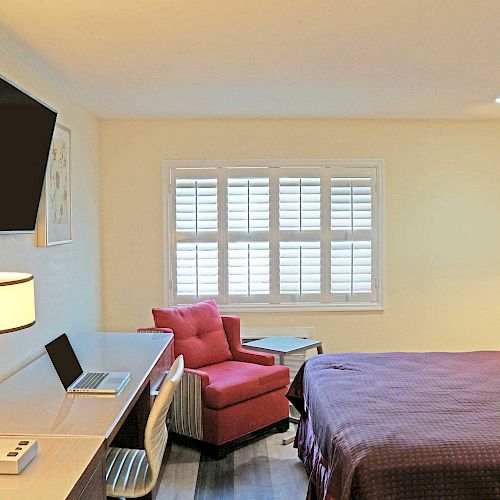 A hotel room with a bed, desk, mounted TV, lamp, red armchair, and window with shutters. Decor includes framed art and minimal furniture.