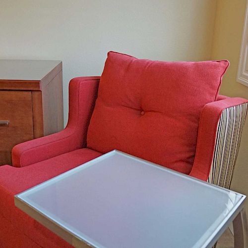 A red chair with a small table attached, a wooden dresser, window shutters, and an air conditioning unit are in the room.