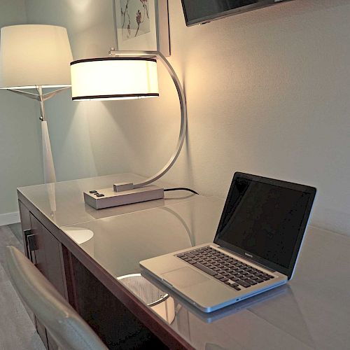 A sleek desk setup with a laptop, modern desk lamp, floor lamp, and a comfortable chair in a well-lit room.