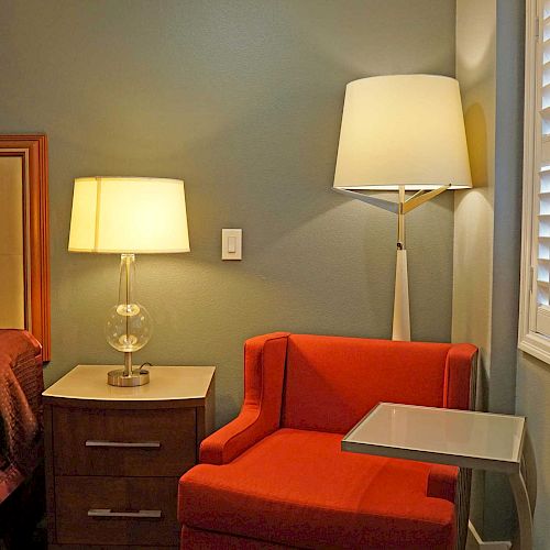 A cozy room with a bed, nightstand, two lamps, a red armchair, a small table, and white shutters on the window.