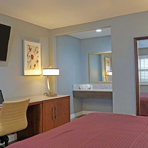 The image shows a hotel room with a bed, a desk with a chair, a TV, a lamp, and a mirror. A bathroom with a sink is visible in the background.