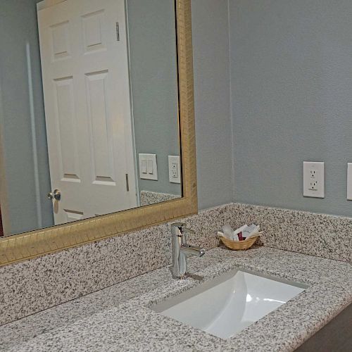 A bathroom vanity with a granite countertop, an under-mount sink, and a large mirror. The background shows a bedroom with a bed and window.