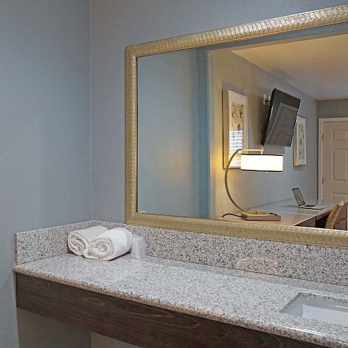 A bathroom countertop with a mirror above, featuring rolled white towels. A TV and a door are reflected in the mirror.