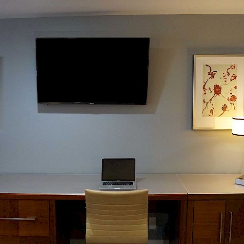 A minimalist workspace with a wooden desk, a laptop, a chair, a table lamp, two framed pictures, and a wall-mounted TV.