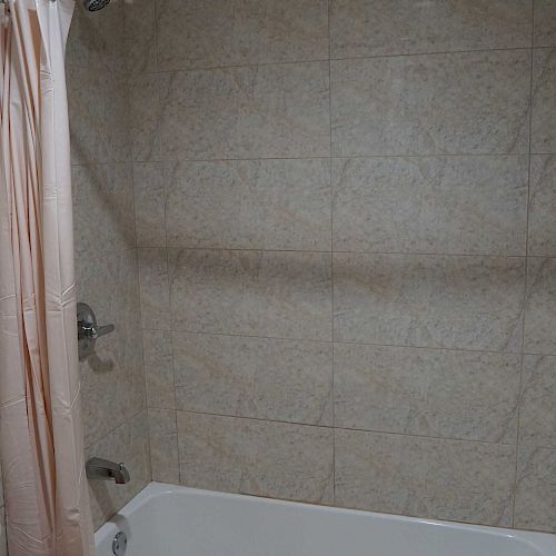 A clean, tiled bathroom with a white bathtub and a pink shower curtain on a curved rod is pictured. The wall tiles are a light beige color.