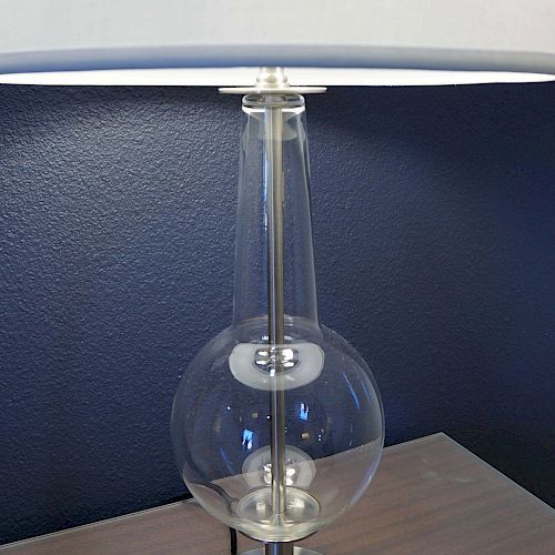 A modern table lamp with a transparent glass base, a metal stand, and a white lampshade, placed on a wooden surface against a dark wall.