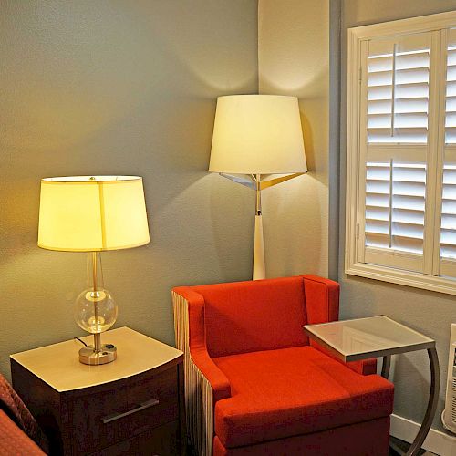 A cozy room corner with a red chair, bedside table with lamp, floor lamp, white shutters, and a wall-mounted air conditioning unit.