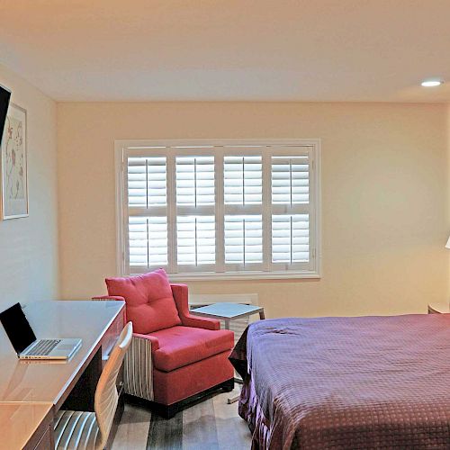 A neatly organized hotel room features a bed, a wall-mounted TV, a desk with a laptop, a red chair, a side table, and a window with shutters.