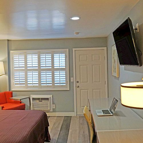 This image shows a hotel room with a bed, desk with a laptop, wall-mounted TV, orange chair, lamps, and artwork on the wall.