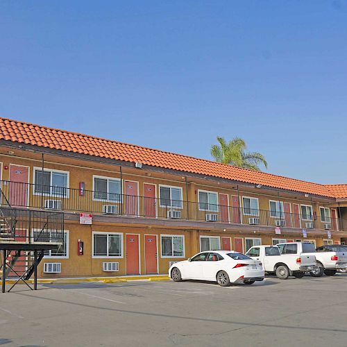 The image shows a two-story motel with a red-tiled roof, exterior staircases, parking lot, and several parked cars, under a clear blue sky.