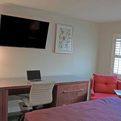 The image shows a modern bedroom with a desk, a lamp, a laptop, an office chair, a wall-mounted TV, framed artwork, a red chair, and a window.