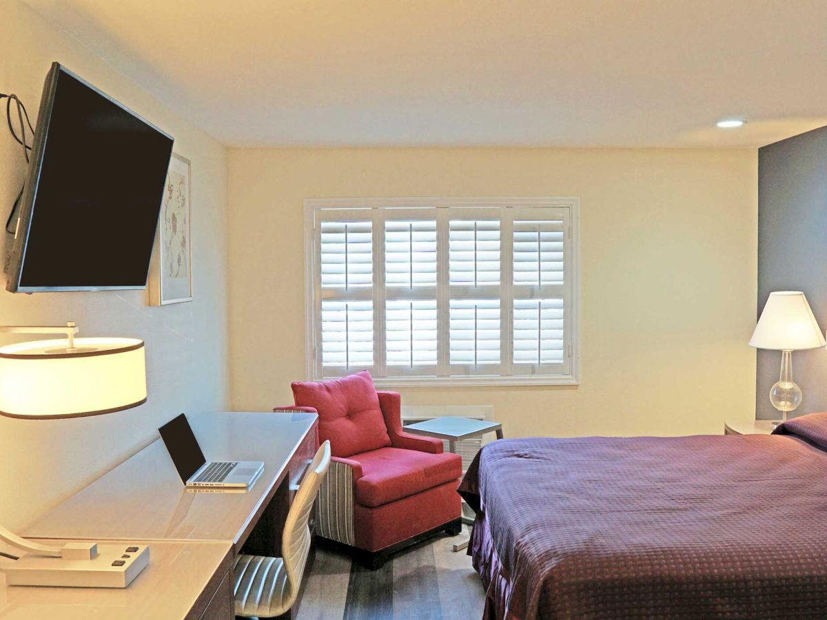 A modern hotel room with a bed, wall-mounted TV, desk, pink chair by the window, and a table lamp with a laptop on the desk.