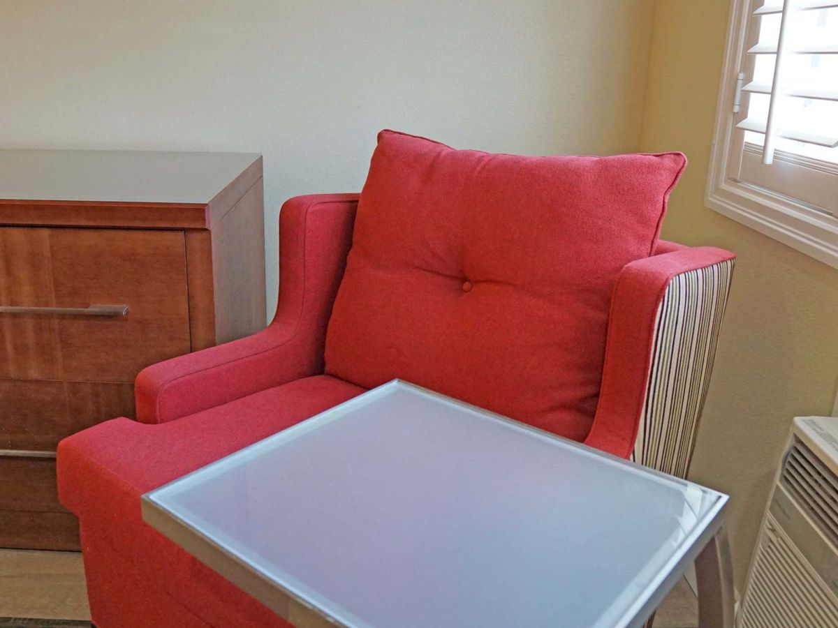 A red armchair with a small table in front, a wooden dresser, and a window with blinds; an air conditioning unit is next to the chair.