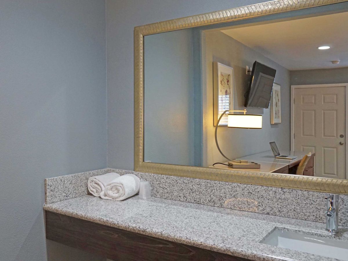 A bathroom with a large mirror, granite countertop, rolled towels, and a sink. The reflection shows a TV, a light fixture, and a door.