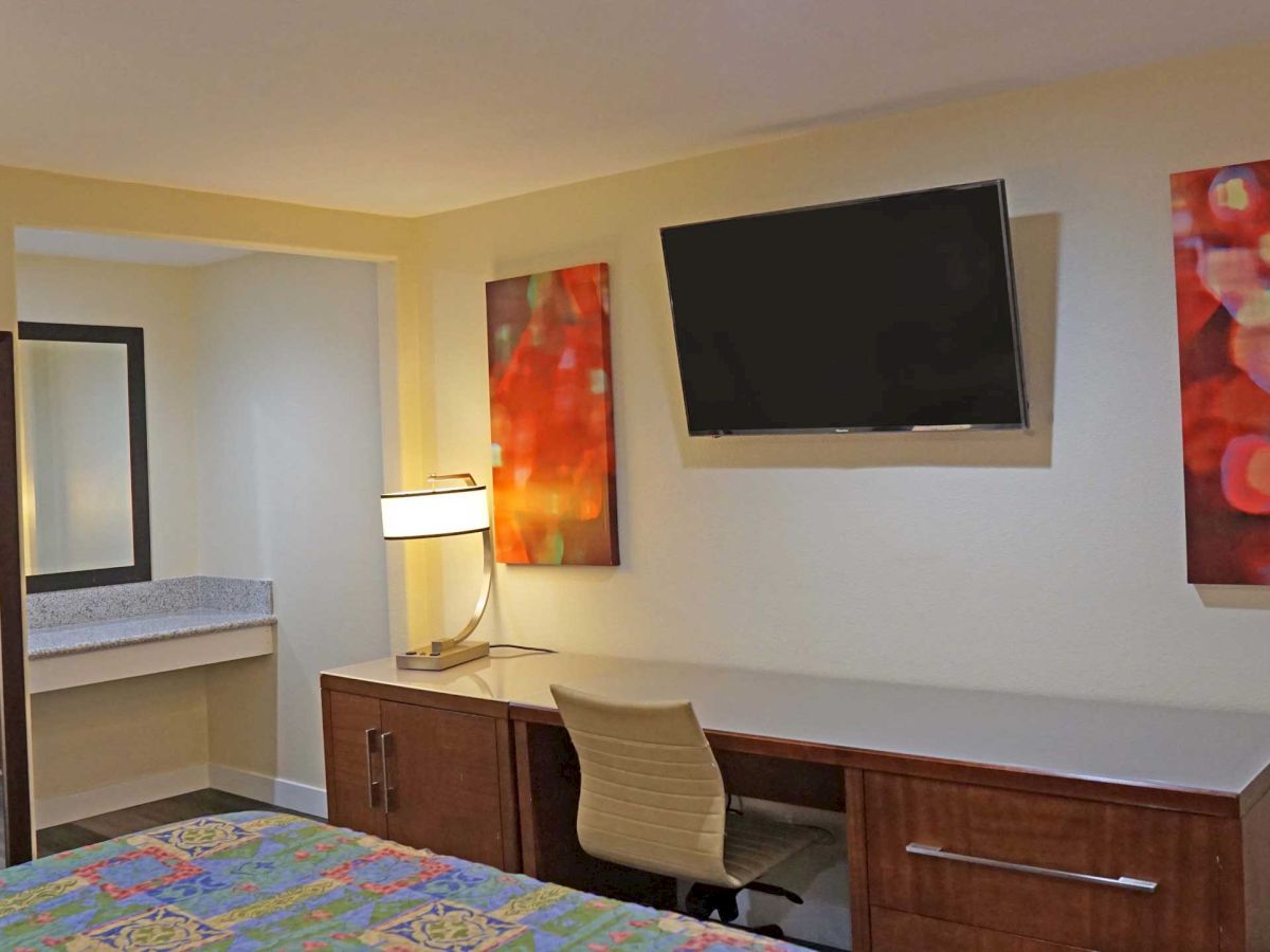 This image shows a hotel room with a wall-mounted TV, a desk with a lamp, two abstract paintings, and a mirror above a counter in the background.