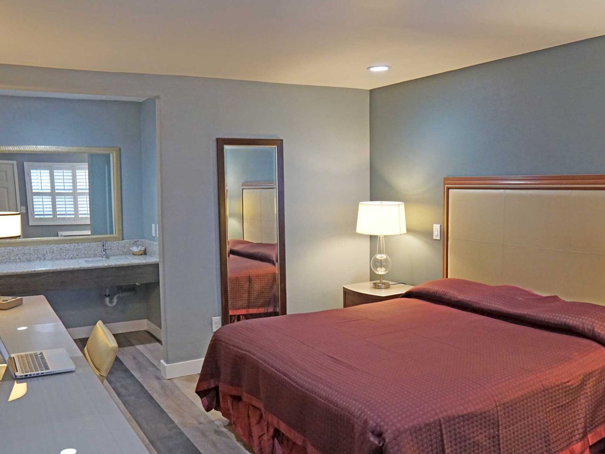 A hotel room with a bed, nightstands, lamps, a mirror, a TV, and a desk with a laptop. Visible bathroom area with a sink and mirror in the background.