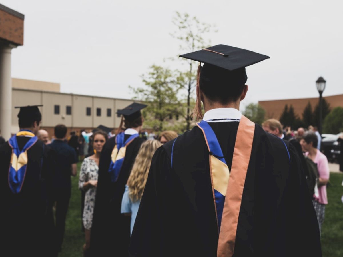 People wearing graduation gowns and caps are gathered outdoors, with others around them. They appear to be celebrating a graduation event.