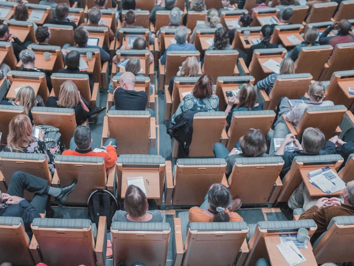 People sitting in rows of seats in an auditorium or lecture hall, viewed from behind, attending an event or presentation.