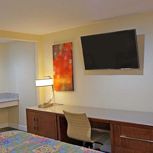 The image shows a modern hotel room with a bed, a TV mounted on the wall, abstract artwork, a desk with a chair, and a lamp on the desk.