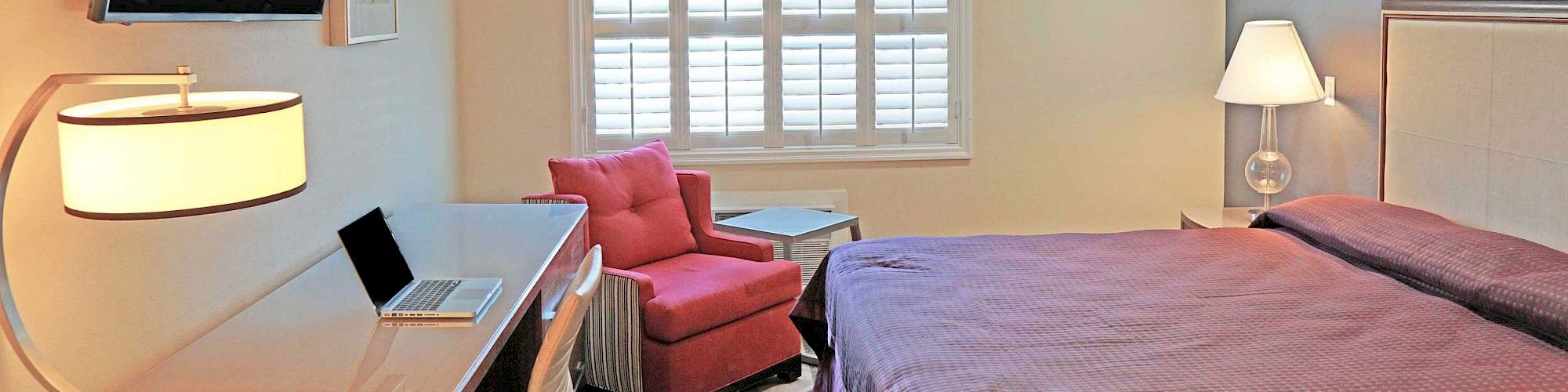 A cozy bedroom features a wall-mounted TV, laptop on a desk, red armchair, table, bed with purple bedding, soft lighting, and shuttered window.